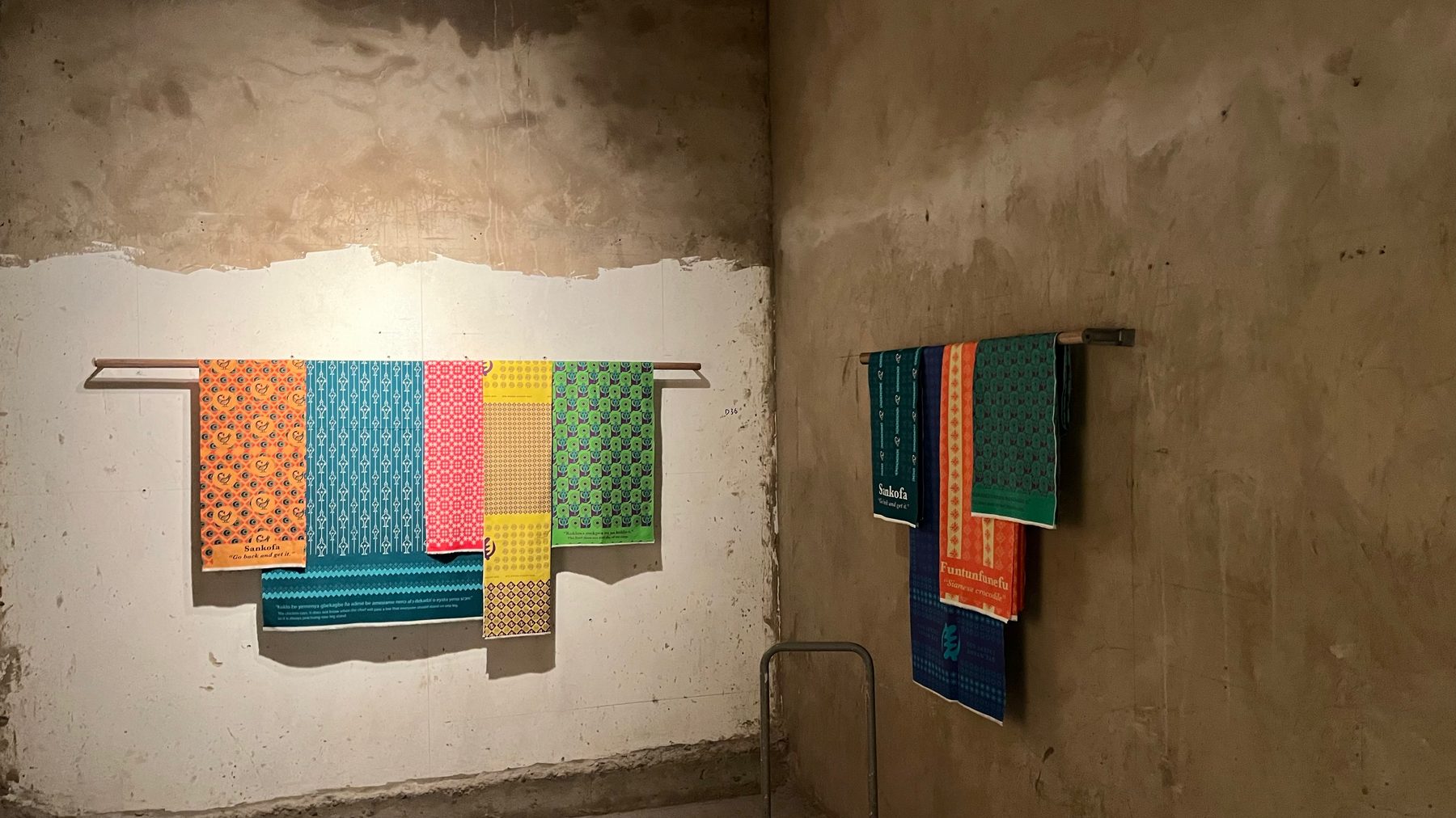 Orange, blue, pink, yellow and green printed cloths hanging from a rail against a whitewashed wall, with darker cloths on the side wall