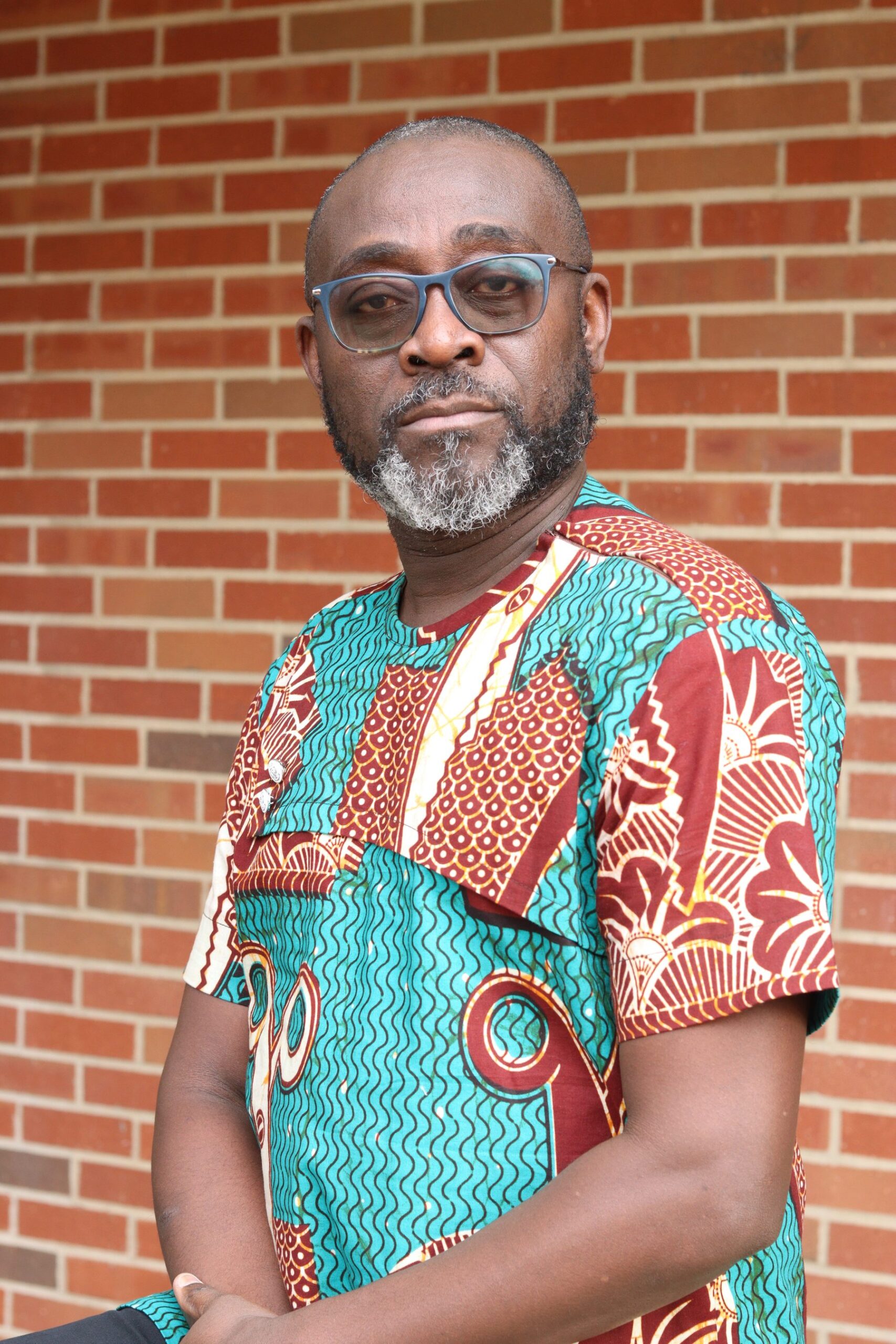 Jasper Dafeamekpor in glasses and a green, red and cream African print shirt, standing in front of a red brick wall
